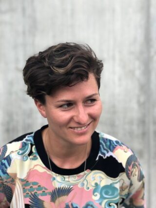 Eine Person mit kurzem, welligem braunem Haar und einem bunt gemusterten Hemd lächelt und blickt vor einem schlichten Hintergrund leicht zur Seite.