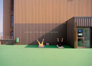 Zwei Personen liegen mit erhobenen Beinen auf Matten vor einer braunen Hauswand mit der Aufschrift „LAUFEN SPRINGEN VERBOTEN“ auf einer grünen Außenfläche unter klarem Himmel.