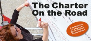 Eine Person schreibt auf eine große Tafel mit dem Text „Die Charta unterwegs“. Ein orangefarbenes Schild weist darauf hin, dass es sich um ein Kunstprojekt von Sylvia Winkler und Stephan Köperl handelt.