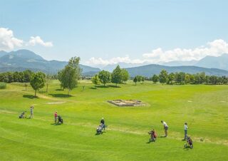 Mehrere Personen spielen Golf auf einem grünen Platz, umgeben von Bäumen und Bergen unter einem klaren, sonnigen Himmel.