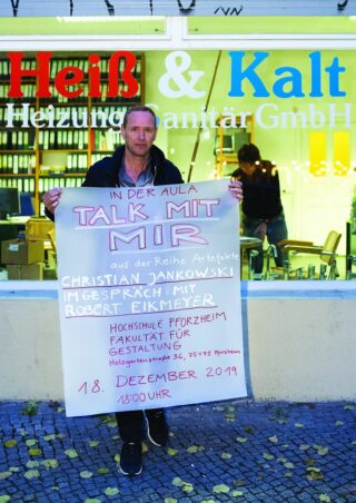 Draußen steht ein Mann mit einem großen handgeschriebenen Schild in deutscher Sprache, das auf eine Vortragsveranstaltung mit Christian Jankowski und Robert Eikmeyer an der Hochschule Pforzheim am 18. Dezember 2019 hinweist.