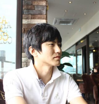 Ein junger Mann mit kurzen schwarzen Haaren und einem weißen Hemd mit Kragen sitzt im Haus neben einer Steinmauer und schaut zur Seite.