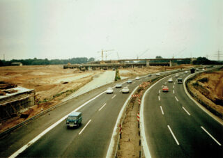 Eine geteilte Autobahn mit mehreren Autos, daneben eine Baustelle mit Kränen und einer unfertigen Überführung im Hintergrund.