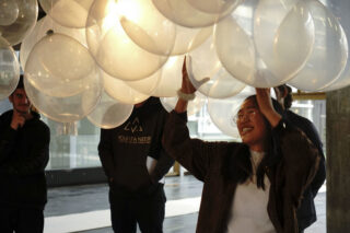 Eine Person lächelt, während sie im Innenbereich eine Gruppe großer transparenter Ballons hochhebt, während andere in der Nähe stehen.