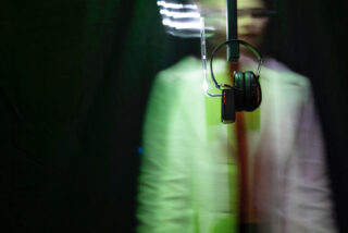 Ein Paar schwarze Kopfhörer hängt im Fokus an einem Ständer, während eine Person in einer weißen Jacke verschwommen vor einem dunklen Hintergrund steht.