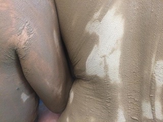 Zwei Menschen mit schlammbedeckter Haut sitzen oder stehen dicht beieinander, Rücken und Arme gegeneinander gedrückt.