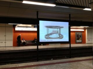 Eine digitale Anzeige auf einem U-Bahnsteig zeigt das Bild einer kreisförmigen Metallstruktur. Drei Personen sitzen auf einer Bank vor einer orangefarbenen Wand, eine vierte Person steht in der Nähe.