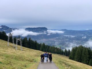 Eine Gruppe von Menschen geht einen gepflasterten Weg auf einem grasbewachsenen Hügel mit Bäumen hinauf und hat unter einem bewölkten Himmel Aussicht auf ein Tal, eine Stadt und Berge.