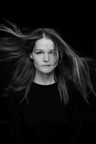 Eine Frau mit glattem Haar und einem schwarzen Oberteil blickt in die Kamera, ihr Haar weht vor einem schwarzen Hintergrund nach außen.