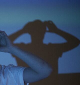 Eine Person in einem weißen Hemd hält ihre Hände an den Kopf und wirft einen Schatten auf eine blaue Wand hinter ihr.