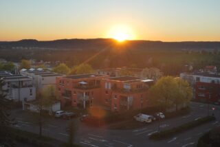 Gezeigt wird ein Wohnviertel mit modernen Gebäuden und geparkten Autos bei Sonnenuntergang, wobei die Sonne hinter fernen Hügeln untergeht.