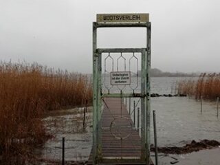 Ein rostiges Metalltor mit der Aufschrift „Bootsverleih“ versperrt den Zugang zu einem überfluteten Holzsteg, der in einen See hineinragt. Ein Schild mit der Aufschrift „Betreten der Zeiten verboten“ ist angebracht.