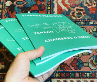 Eine Hand hält drei grüne Bücher mit dem Titel „Strange Teaching Tehran Chambres D’Amis“ mit einer weißen Kartenumrisszeichnung auf dem Einband vor einem gemusterten Teppichhintergrund.