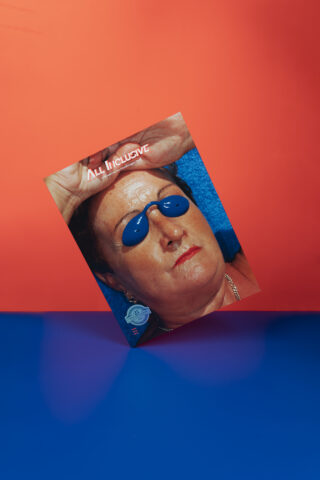 Ein schräges Foto einer Person mit blauer Bräunungsbrille und rotem Lippenstift vor einem blau-roten Hintergrund, mit dem Text „ALL INCLUSIVE“ oben.