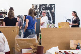 Eine Gruppe von Menschen steht in einem Kunstatelier, einige unterhalten sich, im Hintergrund und Vordergrund sind Kunstwerke und Materialien zu sehen.