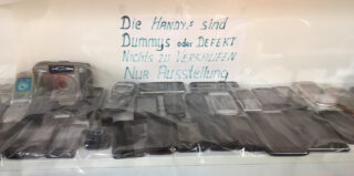 Eine Vitrine mit verschiedenen alten Mobiltelefonen und einem handgeschriebenen Schild auf Deutsch, auf dem steht, dass es sich bei den Telefonen um Attrappen oder defekte Telefone handelt und diese nicht zum Verkauf, sondern nur zur Ausstellung bestimmt sind.