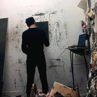 Eine schwarz gekleidete Person steht mit einer Leinwand in der Hand vor einer farbbespritzten Wand; ein Laptop und Künstlerbedarf liegen auf einem kleinen Tisch in der Nähe in einem überfüllten Atelier.