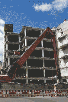 Ein großer Bagger zerstört ein mehrstöckiges Betongebäude, wobei Trümmer herabfallen und davor Sicherheitsbarrieren errichtet werden.