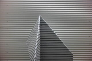 Eine Wellblechstruktur bildet eine spitze Form vor einer Wellblechwand mit horizontalen Linien.