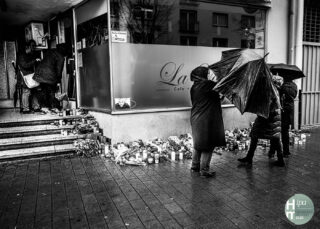 Menschen versammeln sich vor einem Café und legen Blumen, Kerzen und Gedenktafeln auf den nassen Gehweg; manche halten Regenschirme in den Händen. Die Stimmung ist gedrückt.