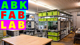 Ein Fertigungslabor mit Arbeitstischen, Lagerregalen mit grünen Behältern, 3D-Druckern und Werkzeugen und einem bunten „ABK FAB LAB“-Schild auf der linken Seite.