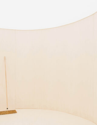 In einem minimalistischen, leeren Raum lehnt ein Holzbesen an einer geschwungenen, hellen Holzwand.