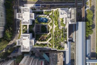 Luftaufnahme eines modernen Gebäudes mit Dachgärten und Grünflächen, umgeben von Straßen, Gehwegen und angrenzenden Gebäuden in einer städtischen Umgebung.