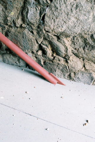 Auf einem hellen Betonboden in der Nähe einer rauen Steinmauer liegt ein kaputtes rotes Rohr, überall sind kleine Trümmer verstreut.