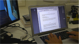 Eine Person tippt auf einer Laptoptastatur, während auf dem Bildschirm ein Dokument geöffnet ist, neben einem Monitor und verschiedenen Schreibtischgegenständen.