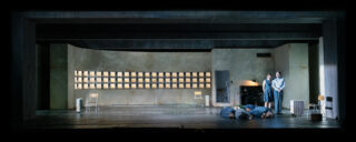 Vier Personen stehen auf der rechten Seite eines kargen, modernen Raumes; drei Personen liegen auf dem Boden, während eine Frau und zwei Männer in der Nähe einer Tür stehen. Drei leere Stühle stehen im Abstand zueinander.