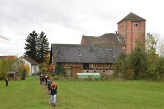 Eine Gruppe von Menschen geht im Gänsemarsch über eine Wiese auf ein großes historisches Gebäude aus Backstein und Holz mit einem Turm und einem Satteldach zu.