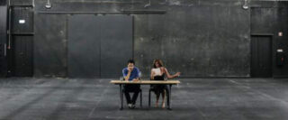 Zwei Personen sitzen mit Getränken an einem Tisch in einem großen, leeren, schwach beleuchteten Raum im Industriestil mit dunklen Wänden und sichtbaren Türen im Hintergrund.