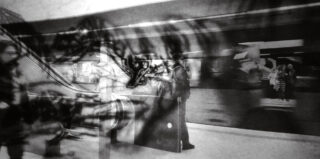 Ein Schwarzweißfoto zeigt Menschen, die in einer U-Bahn-Station stehen und sich bewegen, wobei ein verschwommener Zug und mehrschichtige, abstrakte Überlagerungen einen surrealen Effekt erzeugen.