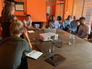 Eine Gruppe von Menschen sitzt mit einem Projektor, Notizblöcken und Glaswaren um einen großen Tisch herum und nimmt an einer Besprechung in einem Raum mit orangefarbenen Wänden teil.