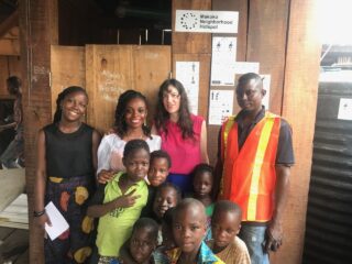 Eine Gruppe von Erwachsenen und Kindern posiert für ein Foto vor einer Holzwand mit einem Schild mit der Aufschrift „Makoko Neighborhood Hotspot“.