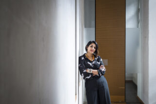 Eine Frau in Geschäftskleidung steht mit verschränkten Armen an einer Wand in einem modernen Büroflur mit großen Fenstern und einer Milchglastür.