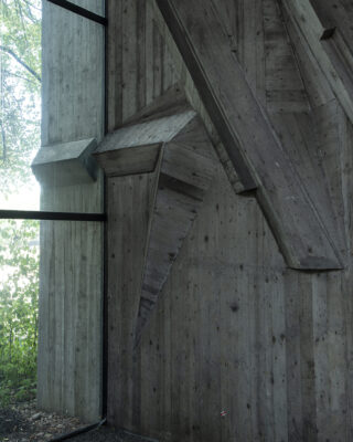 Eckige Betonstruktur mit geometrischen Balken und Paneelen im Inneren eines Gebäudes, neben einem raumhohen Fenster mit sichtbaren Bäumen draußen.