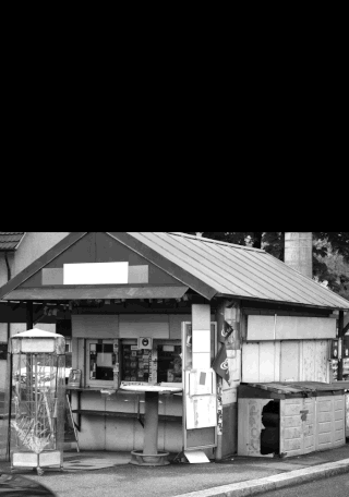 Ein kleiner, verwitterter Kiosk mit einer Telefonzelle auf der linken Seite, draußen sind auf Regalen verschiedene Plakate und Gegenstände ausgestellt; Foto in Schwarzweiß.