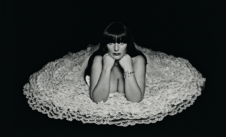 Eine Frau mit dunklem Haar und Pony stützt ihr Kinn auf ihre Hände und liegt auf einem runden, gerüschten Spitzengewand vor schwarzem Hintergrund.