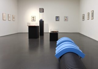 In einem Kunstgalerieraum sind gerahmte Kunstwerke an weißen Wänden sowie verschiedene Skulpturen und Objekte auf Sockeln ausgestellt; im Vordergrund steht eine blau-schwarze zylindrische Struktur auf dem Boden.