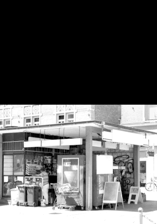 Eine Person steht neben Einkaufswagen und Mülltonnen vor einem kleinen städtischen Markt, im Hintergrund sind Graffiti an der Wand zu sehen.