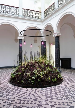 Im Innenbereich ist ein runder Erdhügel mit jungen Pflanzen ausgestellt, umgeben von Marmorsäulen und violetten Hängelampen in einem Atrium mit gemusterten Fliesen.