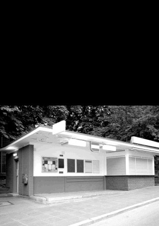 Ein kleiner, geschlossener Kiosk mit leeren Schildern und Fensterläden steht auf einem gepflasterten Platz, umgeben von Bäumen. Das Bild ist schwarz-weiß.