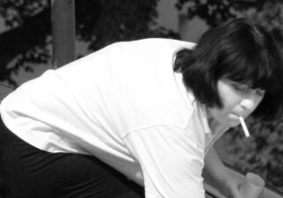 Eine Frau mit dunklem Haar und hellem Hemd hält eine Zigarette im Mund und eine Tasse in der Hand.