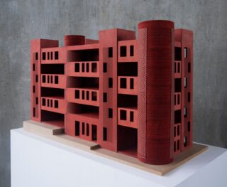 Ein kleines Architekturmodell eines modernen, rechteckigen roten Gebäudes mit zylindrischen Eckstrukturen, das auf einem weißen Sockel vor einer grauen Betonwand steht.