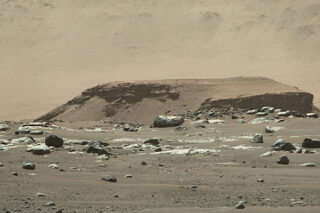 Felsiges, staubbedecktes Gelände mit verstreuten Felsbrocken und einem flachen Hügel unter einem dunstigen Himmel, das an eine Marslandschaft erinnert.