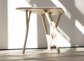 Ein heller Holztisch mit drei unregelmäßigen, astartigen Beinen steht auf einem gefliesten Boden vor einer weißen Wand mit diagonalen Schatten.