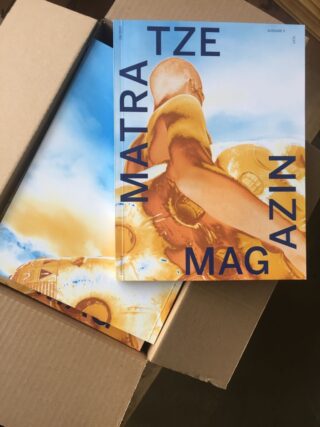 In einem offenen Karton befinden sich Exemplare einer Zeitschrift mit dem Titelbild einer Person in Gelb und der fettgedruckten blauen Schrift „MATRATZE MAGAZIN“.