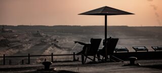 Zwei Menschen sitzen unter einem großen Sonnenschirm und blicken bei Sonnenuntergang auf einen Tagebau. In der Ferne sind Industriemaschinen zu sehen.