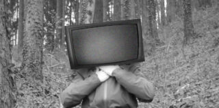 Eine Person steht im Wald. Anstelle ihres Kopfes zeigt ein alter Fernseher statisches Rauschen. Das Bild ist schwarz-weiß.